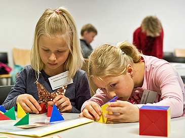 Two girls sit at a table and work with colorful geometric shapes. In the background you can see other children.