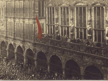 Menschen hissen vor Zuschauern eine rote Fahne auf dem Balkon eines Rathauses.