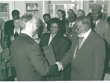 Black and white photo. Assembly with men and women. In the foreground two men shake hands